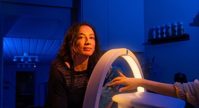 Person sitter vid ett manikyrbord medan en hand i blå engångshandske arbetar under en rund skrivbordslampa i en upplyst salong.